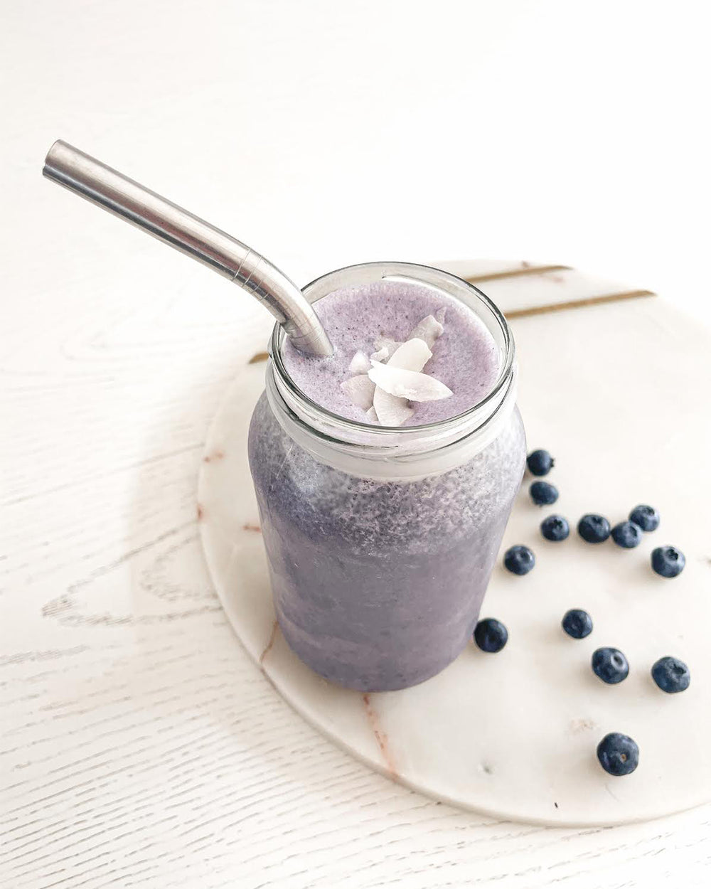 Image of a glass of purple smoothie with blueberries scattered on the table.