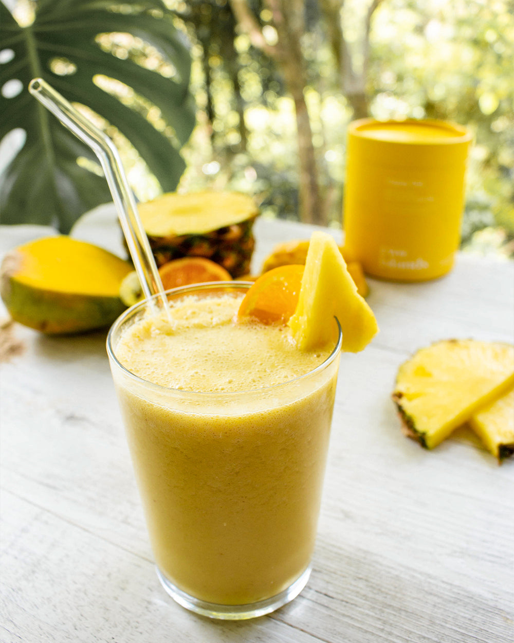 Image of a yellow smoothie in a glass with a glass straw with pineapples, mangoes and Happy Gut tub at the background.